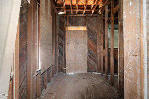 A small, narrow room with exposed wooden framing and a partially covered window. The walls show wood paneling, and the floor is bare with scattered debris. The ceiling has visible wooden rafters. A door leads to another area.