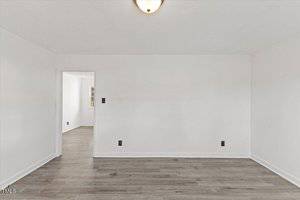An empty room with white walls and a wooden floor. There's a doorway leading to another room in the background. A ceiling light is visible, and electrical outlets are installed on the walls.