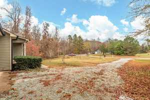 A rural outdoor scene featuring a gravel driveway leading to a house partially visible on the left. The area is surrounded by trees with autumn foliage. A few parked vehicles are seen in the distance under a partly cloudy sky.