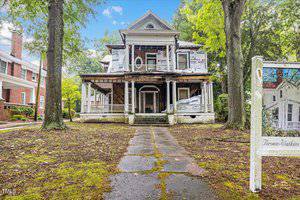 A large, partially renovated two-story house with a wraparound porch and white siding stands at 217 College Street in Oxford. The front yard is unkempt, with patches of moss on the pathway. A sign displaying architectural drawings is visible on the right, while tall trees gracefully surround the property. .