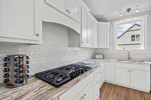 A modern kitchen features white cabinets and a marble countertop. A gas stove is built into the counter with a spice rack nearby. A large window above the sink allows natural light to brighten the space. The backsplash is made of light-colored tiles.
