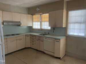 A dimly lit kitchen features beige cabinets, a white countertop, a built-in stove, and a range hood. A dishwasher is visible beneath the counter. Two windows with blinds allow limited natural light. The floor appears to be linoleum.