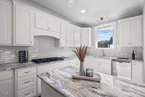 Modern kitchen with white cabinets and marble countertops. A large window provides natural light. The island features a decorative tray with a vase of dried plants and candles. Stainless steel appliances and a tile backsplash complete the space.