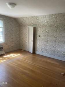 An empty room with floral wallpaper and a wooden floor. There is a single window on the left, allowing sunlight to enter the space. A closed white door is centered on the patterned wall.