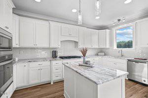 A modern kitchen with white cabinets, a marble countertop island, stainless steel appliances, and a gas stove. The space is illuminated by pendant and recessed lighting. A window offers a view of trees outside. There is a vase with dried flowers on the island.