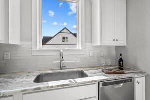 A kitchen with white cabinets and a marble countertop features a sink, dishwasher, and cutting board with a bottle of wine and two glasses. A window above the sink shows an outdoor view of a house and blue sky with clouds.