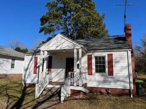 A small, single-story house with white siding and a gray roof, featuring a front porch with steps and red shutters on the windows. There is a chimney on the right side and a large tree in the background under a clear blue sky.