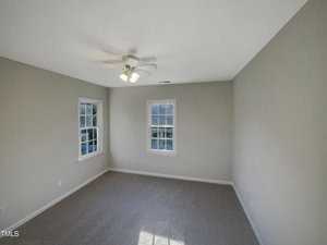 A vacant room with beige walls and a gray carpeted floor. There is a ceiling fan with lights, and three windows providing natural light.