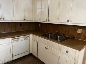 A kitchen with worn white cabinets, a brown countertop, and a double stainless steel sink. A white dishwasher is visible beneath the counter. The walls have a wood-paneled backsplash, and there's an electrical outlet on the right.