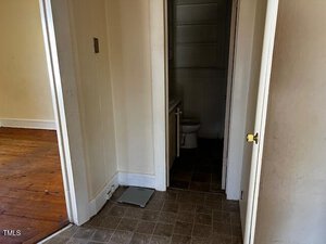 A view of a small bathroom with a visible toilet, adjacent to a room with wooden flooring. The open doorway reveals tile flooring leading into the bathroom. A scale is on the floor near the entrance.