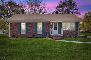A single-story brick house at 403 Person Street, with green shutters, is set against a vibrant pink and purple sunset. This Oxford charmer features a front door with a small porch and white railing, surrounded by a grassy lawn and trees in the background.