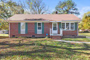 A single-story red brick house with a brown shingle roof, white trim, and blue shutters. A white front door is centered between windows. Surrounding the house is a grassy yard with scattered leaves and trees in the background.