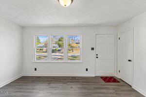 Room interior with light gray wood flooring, white walls, and a ceiling light. There are three large windows and a white door with black hardware. A small red rug is placed near the door. Outside, trees and a street are visible.
