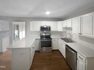 A modern kitchen with white cabinets and gray countertops. Features stainless steel appliances, including a refrigerator, stove, microwave, and dishwasher. Hardwood flooring is visible, and there is a windowed door leading outside.