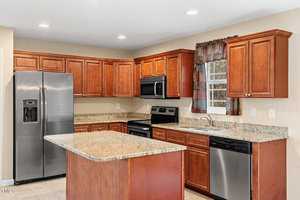 A modern kitchen features wooden cabinets, a marble island countertop, and stainless steel appliances, including a refrigerator, stove, microwave, and dishwasher. A window with patterned curtains is above the sink, and recessed lighting brightens the space.