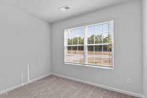 An empty room with white walls and a large window featuring blinds. The floor is carpeted in a neutral tone, and natural light is streaming through the window, casting shadows on the walls. The view outside shows trees and a paved surface.