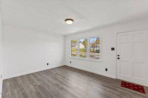 An empty room with white walls and a gray wooden floor features three windows and a white door. A ceiling light fixture is centered on the ceiling. A red doormat is placed at the entrance. Outside, a residential neighborhood is visible.