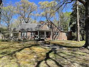 A brick house at 710 Hillboro Street, with a gabled roof, is surrounded by tall trees in sunny Oxford. The front yard features a wooden fence, casting shadows from the branches onto the ground. Above, the sky is clear and blue.