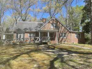 A brick house with a slanted roof, a small chimney, and multiple windows. A wooden ramp leads to the front entrance. The house is surrounded by trees, and the yard appears grassy with patches of sunlight.