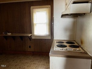 A small kitchen area with wood panel walls and a tiled floor. The room features a white stovetop with four burners and an exhaust hood above. A single window with blinds is on one wall, beside a narrow shelf. The room appears dimly lit.