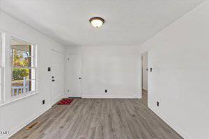 An empty room with white walls and a light wood floor. There are two visible doors, one with a window and another leading to another room. A ceiling light fixture is present, and a small red mat lies near the door with the window.