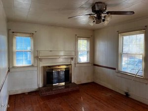A small, empty room with wooden floors and white paneled walls. It features a brick fireplace with a wooden mantel, two windows with blinds, and a ceiling fan with lights. The room appears well-lit from natural light.