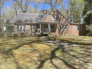 A red brick house with a gabled roof stands surrounded by tall trees. It features a small front porch with a wooden ramp, several windows, and a chimney. The foreground shows a patchy grassy area and some shadows from the trees.