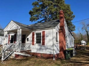A small, single-story white house with red shutters and a brick chimney. There is a front porch with steps leading up, a few trees nearby, and a green garbage bin at the side. The sky is clear and blue.