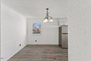 A small, empty room with white walls and wood-look flooring. A window shows an outdoor view. A simple ceiling light fixture with three shades hangs from the ceiling. A stainless steel refrigerator is partially visible on the right.