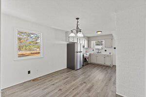 A bright kitchen with white cabinets and walls, a stainless steel refrigerator, and a stove. A black chandelier hangs from the ceiling above a wood-look floor. A window on the left and a view of a backyard with trees are visible.