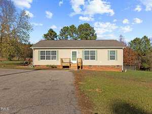 A single-story house with beige siding and a brown shingled roof, located at 4509 NC Hwy 56 in Franklinton. It features a small front porch with steps leading to the entrance, set on a large paved area surrounded by grass and trees under a partly cloudy sky.