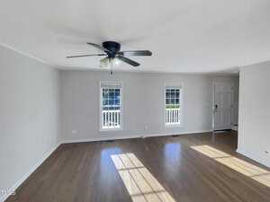 An empty room features polished hardwood floors and white walls. A ceiling fan with lights is centered above. Two windows and a door let in natural light, casting shadows on the floor.