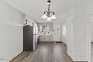 A kitchen with white walls and dark wood flooring features a stainless steel refrigerator and a sink beneath a window. There are cabinets above and below the counter. A chandelier hangs from the ceiling, and a door leads outside.