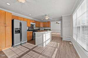 A kitchen with wooden cabinets and a dark countertop. There is a stainless steel refrigerator, an electric stove, and a microwave. An island with a sink is in the center. The flooring is wooden, and large windows bring in natural light.