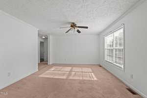 A minimalist living room with plain white walls and beige carpet. A ceiling fan with brown blades is centrally mounted. Sunlight streams through a window with blinds, casting shadows on the carpet. An open doorway leads to another room.