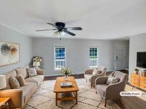 A virtually staged living room features a ceiling fan, two windows, and a door. The room includes a sofa, two armchairs, a coffee table, and a sideboard. Decor items such as a rug, plants, and wall art are present.