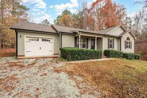 A single-story house with green siding, a one-car garage, and a front porch is surrounded by autumnal trees. The lawn is partially covered with dry leaves, and a gravel driveway leads to the garage. The sky is partly cloudy.