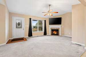 A living room with beige walls and carpet, featuring a ceiling fan and a window with black curtains. A fireplace under a wall-mounted TV is on the right. A white door with a decorative glass panel is on the left. The room is well-lit and empty.
