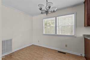 An empty room with light-colored walls and wooden flooring. There is a three-light chandelier on the ceiling. Two windows with blinds allow natural light in. A vent is on the lower wall next to a wooden cabinet on the right.