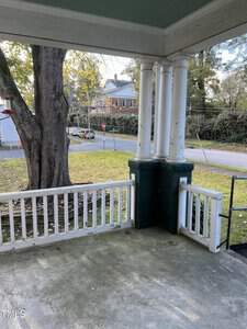 A view of a porch featuring white columns and a railing. The porch overlooks a quiet suburban street with a large tree to the left and a brick building in the background. The ground is covered with fallen leaves.