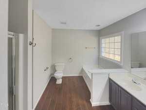 A bathroom with a toilet, a bathtub, and a glass-enclosed shower. There is a large window above the bathtub, a long countertop with a sink, and wooden flooring. The walls are painted in a light color.