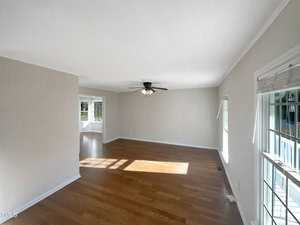 An empty living room with wooden floors and neutral-colored walls. A ceiling fan is centered above. Sunlight streams through multiple windows, creating patterns on the floor. A small, adjacent area is visible, separated by a corner wall.