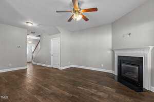 A spacious living room with wooden flooring, a ceiling fan with lights, and an empty wall mount for a television above a white-trimmed fireplace. A white door leads to a hallway with stairs visible in the background.