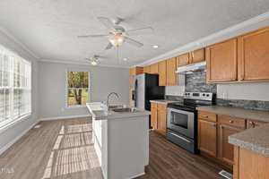 A modern kitchen with wooden cabinets, a central island with a sink, and stainless steel appliances, including a stove and refrigerator. There are two ceiling fans, large windows, and wood flooring.