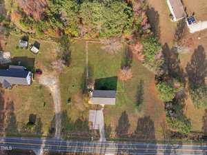 Aerial view of a rural scene with a single-story building surrounded by grass and trees. A driveway leads to the building, and adjacent properties are visible. A road runs horizontally at the bottom of the image.