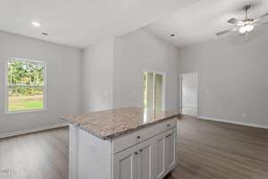 Interior view of a modern, unfurnished kitchen and living area with light gray walls and wood flooring. A granite kitchen island is centered, and a ceiling fan is visible in the living space. A window and a glass door show an outdoor view.