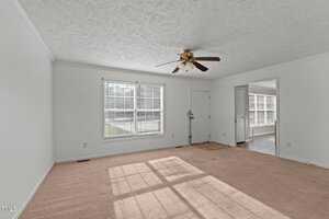 A bright, empty living room with a beige carpet and textured white ceiling. A ceiling fan is centered above. A large window and a door are on one wall. An open doorway leads to another room with additional windows and light wood flooring.