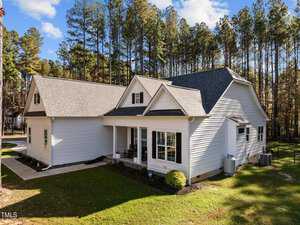 A modern, single-story white house with a dark gray gabled roof is shown on 4321 S Greg Allen Way. The house features multiple windows and a covered porch, set amid tall pine trees in Oxford under a partly cloudy sky.