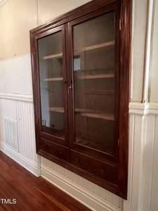 A dark wood built-in cabinet with glass doors and two drawers is set in a white paneled wall. The cabinet has empty shelves and a polished wooden floor in front of it.