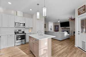 Modern kitchen and living area with wooden floors. The kitchen features white cabinets, a stainless steel stove, and a central island with pendant lights. The living area has a gray sectional sofa, a wall-mounted TV, and a deer head mounted above a fireplace.